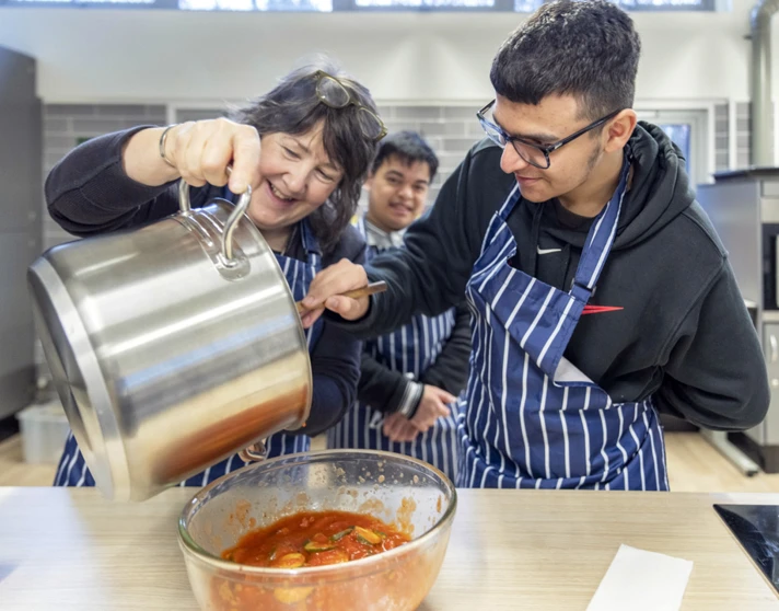 Woman pouring sauce from a large pot into a bowl while smiling and working with two young men in striped aprons. Woman pouring sauce from a large pot into a bowl while smiling and working with two young men in striped aprons.